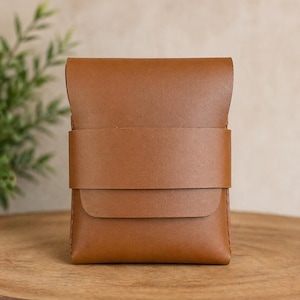 May include: A small, rectangular, brown leather wallet with a flap closure and a wide band detail. The wallet is sitting on a wooden surface, with a blurred green plant in the background.