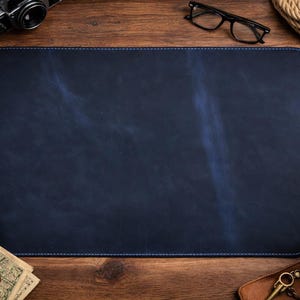 May include: A navy blue leather desk mat with blue stitching. The mat is rectangular and sits on a wooden surface. Other items on the desk include a camera, glasses, a pen, a compass, a cup of coffee, and a wallet.