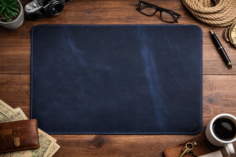 May include: A navy blue leather desk mat with blue stitching. The mat is rectangular and sits on a wooden surface. Other items on the desk include a camera, glasses, a pen, a compass, a cup of coffee, and a wallet.