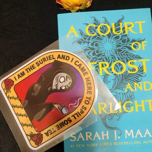 May include: A Court of Frost and Starlight book with a sticker that reads "I am the Suriel and I came here to spill some tea." The book is light blue with yellow text and a floral design. A dried yellow rose is in the background.
