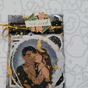 May include: A black mesh gift bag with gold accents and a gold drawstring. The bag contains stickers with a couple embracing under a crescent moon and stars. A pin with the words "book nerd" is attached to the bag.