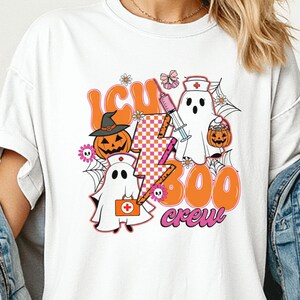 Puede incluir: Una camiseta blanca de manga corta con un estampado gráfico que incluye el texto ICU BOO crew. El diseño muestra dos fantasmas con gorros de enfermería, una calabaza con sombrero de bruja, una jeringuilla, un rayo a cuadros y telarañas.