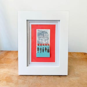 May include: A framed artwork featuring a black and white drawing of a building with a red background. A row of blue matchsticks with black fish silhouettes are placed below the drawing.