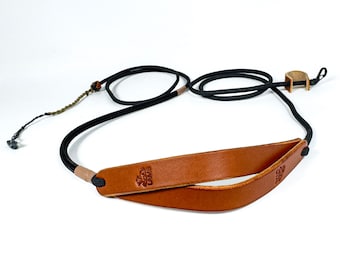 May include: A camera strap featuring a tan leather neck pad embossed with the word LAMB and a logo, connected to a black braided cord. The strap includes light tan leather accents, a small metal ring, and a thin cord extension with a metal tip.