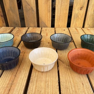 May include: A collection of seven small, round ceramic bowls in various colors, including blue, brown, white, and orange. The bowls have a ribbed design and are arranged on a wooden surface.