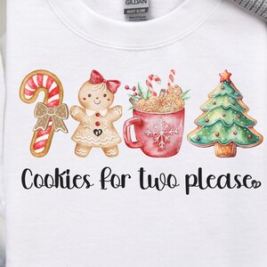 Könnte beinhalten: Weißes Sweatshirt mit festlichem Design mit Zuckerstange, Lebkuchenmann, Tasse mit Keksen und Weihnachtsbaum. Der Text "Cookies for two please" ist unter den Illustrationen aufgedruckt. Eine graue Mütze und Weihnachtsdekoration sind ebenfalls zu sehen.