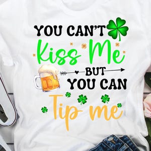 May include: White t-shirt with the text "You Can't Kiss Me But You Can Tip Me" in green, black, and yellow. The design includes a beer mug and shamrocks. A pair of white sneakers and blue jeans are visible.