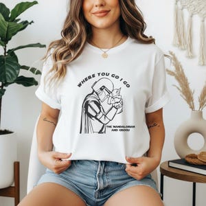 Puede incluir: Camiseta blanca con un diseño en blanco y negro de The Mandalorian sosteniendo a Grogu, con el texto "WHERE YOU GO I GO" arriba y "THE MANDALORIAN AND GROGU" abajo. Cuello redondo.