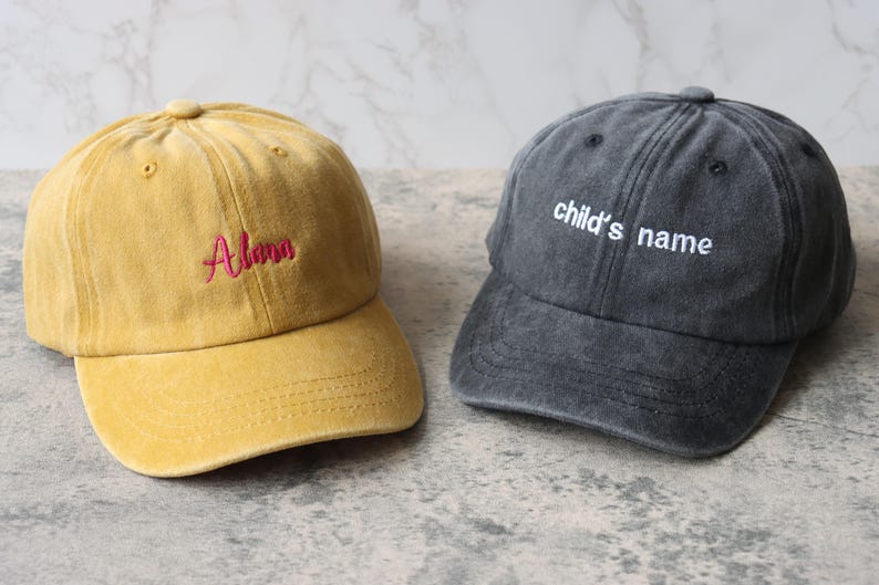 Puede incluir: Dos gorras de b&eacute;isbol, una amarilla con el nombre "Alana" bordado en rojo, y una gris con el texto "child's name" bordado en blanco.