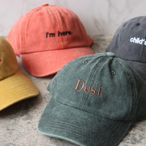 Puede incluir: Cuatro gorras de b&eacute;isbol de diferentes colores con texto bordado. Las gorras son amarillas, naranjas, verdes y negras. El texto en las gorras dice "Always", "I'm here", "Desi" y "child's name".
