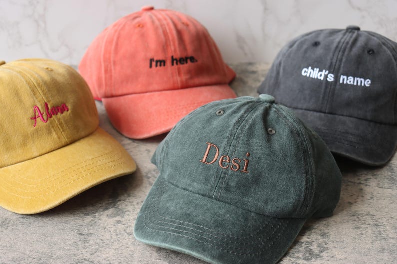 Puede incluir: Cuatro gorras de b&eacute;isbol de diferentes colores con texto bordado. Las gorras son amarillas, naranjas, verdes y negras. El texto en las gorras dice "Always", "I'm here", "Desi" y "child's name".