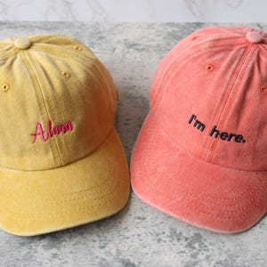 Puede incluir: Dos gorras de b&eacute;isbol, una amarilla con el nombre "Alana" bordado en rosa, y la otra naranja con la frase "Estoy aqu&iacute;" bordada en negro.