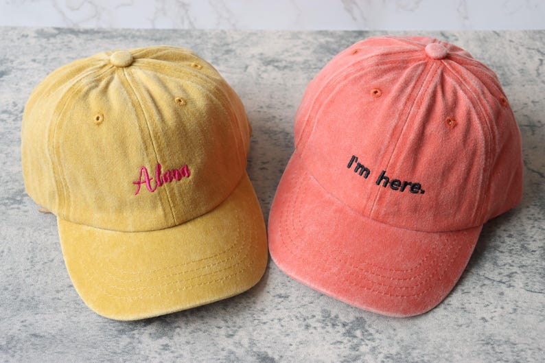 Puede incluir: Dos gorras de b&eacute;isbol, una amarilla con el nombre "Alana" bordado en rosa, y la otra naranja con la frase "Estoy aqu&iacute;" bordada en negro.