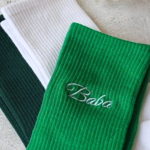 May include: A close-up shot of three pairs of ribbed socks in green, white, and dark green. The green sock features the embroidered word "Baba" in a cursive font. The socks are folded, highlighting their texture and colours.