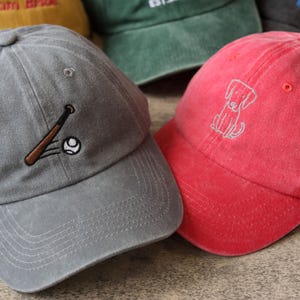 Pode incluir: Tr&ecirc;s bon&eacute;s de beisebol com diferentes desenhos bordados. Um bon&eacute; &eacute; cinza com um desenho de beisebol e taco, outro &eacute; vermelho com um desenho de cachorro e o &uacute;ltimo &eacute; verde com a palavra "EST." bordada.