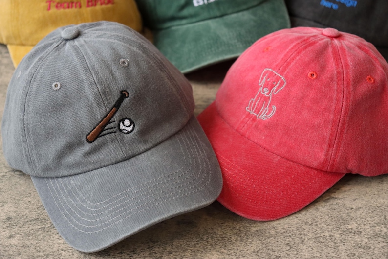 Pode incluir: Tr&ecirc;s bon&eacute;s de beisebol com diferentes desenhos bordados. Um bon&eacute; &eacute; cinza com um desenho de beisebol e taco, outro &eacute; vermelho com um desenho de cachorro e o &uacute;ltimo &eacute; verde com a palavra "EST." bordada.