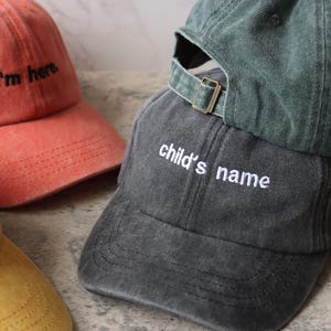 Puede incluir: Tres gorras de b&eacute;isbol de diferentes colores con texto bordado. La primera gorra es naranja con el texto "I'm here". La segunda gorra es verde oscuro con una correa blanca y el texto "child's name". La tercera gorra es amarilla con el texto "Mom".