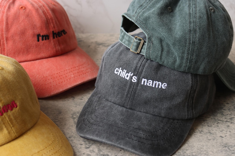 Puede incluir: Tres gorras de b&eacute;isbol de diferentes colores con texto bordado. La primera gorra es naranja con el texto "I'm here". La segunda gorra es verde oscuro con una correa blanca y el texto "child's name". La tercera gorra es amarilla con el texto "Mom".