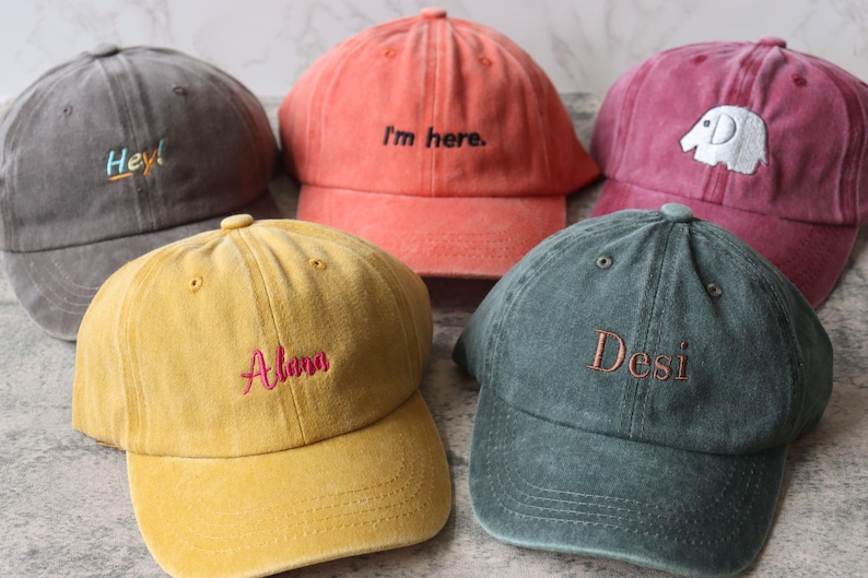 Puede incluir: Cinco gorras de b&eacute;isbol de diferentes colores con texto bordado. Las gorras son grises, rojas, amarillas, verdes y naranjas. El texto en las gorras dice "Hey!", "I'm here", "Aloha", "Desi", y un gr&aacute;fico de elefante.