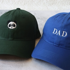Peut inclure: Deux casquettes de baseball, l'une verte avec un panda noir et blanc brodé sur le devant et l'autre bleue avec le mot "DAD" brodé en blanc sur le devant.