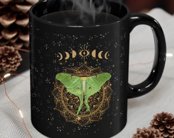 May include: A black ceramic mug featuring a vibrant green luna moth centered over a detailed gold mandala design. Above the moth, a series of gold moon phases and a central eye symbol are displayed against a dark background with scattered stars.