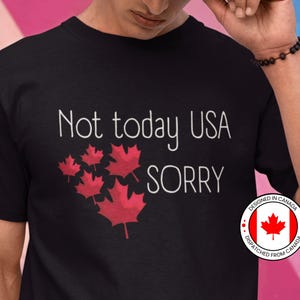 May include: Black t-shirt with the text "Not today USA sorry" in white. The shirt features a cluster of red maple leaves. A circular sticker with the text "Designed in Canada" and a red maple leaf on a white background is on the right side of the shirt.