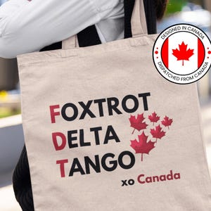 May include: A beige tote bag with the words "FOXTROT DELTA TANGO" in black letters with red outlines. The bag also has a red maple leaf and the text "DESIGNED IN CANADA DISPATCHED FROM CANADA" in a circle. The bag also has the text "xo Canada" in black letters.