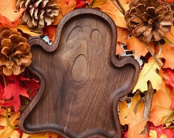 May include: A wooden ghost-shaped serving tray with a carved face. The tray is brown and has a smooth finish. It is surrounded by fall leaves.