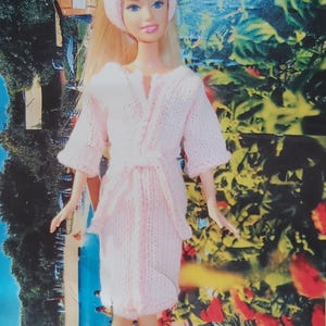 Pode incluir: Uma boneca loira veste um roupão de malha rosa claro com uma faixa de cabelo a combinar. O roupão apresenta um cinto para atar na cintura, mangas a três quartos e um padrão de malha texturizado. A boneca tem olhos azuis e expressão neutra.