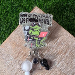 May include: A badge reel with a frog illustration using a laptop, with the text "NONE OF YOUR EMAILS ARE FINDING ME WELL". The frog is green, and the laptop screen is pink. The reel has a white and black beaded accent.
