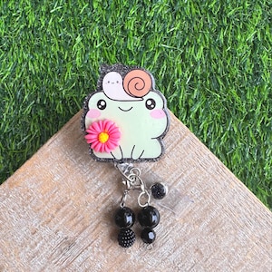 May include: A light green frog-shaped badge reel with a snail and pink flower design. The badge reel has a black glitter border and black beaded accents. The reel is on a wooden surface with a green background.