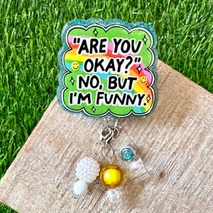 May include: A badge reel with the text "ARE YOU OKAY?" and "NO, BUT I'M FUNNY." The badge has a green border and a rainbow-colored background. It is decorated with a white, yellow, and clear bead charm.
