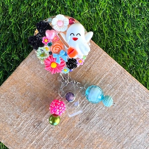 May include: A circular badge reel decorated with a white ghost figurine holding an orange broom, surrounded by colorful miniature flowers, hearts, a blue bow, and black accents. Two dangling charms feature pink, green, and blue beads attached to the clip.