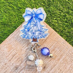 May include: A Christmas tree-shaped badge reel with a blue bow and floral design. The badge reel has a clear plastic holder, a silver clip, and a charm with blue and iridescent beads. The tree is approximately 3 inches tall.