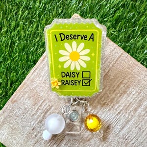 May include: A green badge reel with the text "I Deserve A Daisy Raisey". The badge features a daisy graphic and a checkmark. The badge reel has a clear plastic base with decorative beads.