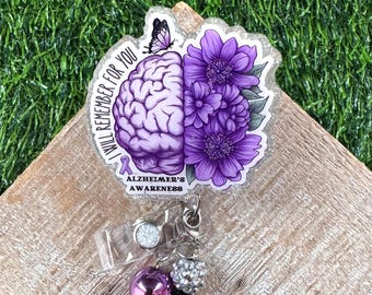 May include: A purple and white Alzheimer's Awareness badge reel. The design features a brain, flowers, and a butterfly. Text on the badge reads "I will remember for you" and "Alzheimer's Awareness". The reel has a clear plastic clip and decorative beads.