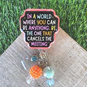 May include: A badge reel featuring a black, scalloped-edge acrylic topper with glittery pink borders. The text reads: IN A WORLD WHERE YOU CAN BE ANYTHING, BE THE ONE THAT CANCELS THE MEETING. It includes a silver clip with orange and blue beads.