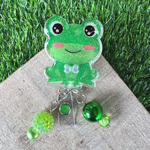May include: A green glittery frog badge reel featuring a small white bow tie, large black eyes with white highlights, and pink cheeks. Two silver chains hang from the bottom, each adorned with green beads in varying textures and shades.