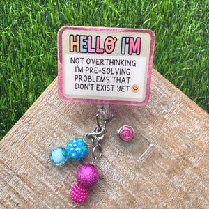 May include: A pink and white badge reel with the text "HELLO I'M NOT OVERTHINKING I'M PRE-SOLVING PROBLEMS THAT DON'T EXIST YET" and a smiley face. The badge reel has a clear plastic clip and a charm with blue and pink beads.