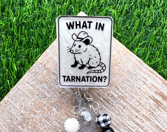May include: A rectangular badge reel featuring a white background with a black line drawing of an opossum wearing a cowboy hat. The text reads WHAT IN TARNATION? The reel includes a clear plastic strap and two dangling charms with beads.