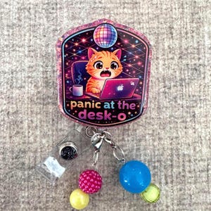 May include: A badge reel with a cat graphic, a laptop, and the text "panic at the desk-o." The badge reel has a clear plastic clip, a black glitter accent, and colorful beads. The cat is orange and white with wide eyes.