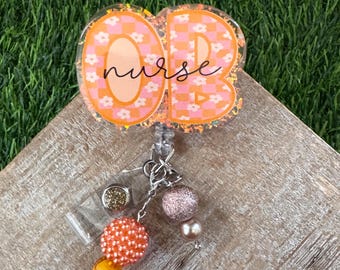 May include: A badge reel with the word "nurse" in cursive script. The badge reel has an orange and white floral design with a glittery border. The reel is attached to a beaded charm with orange, pink, and gold beads.