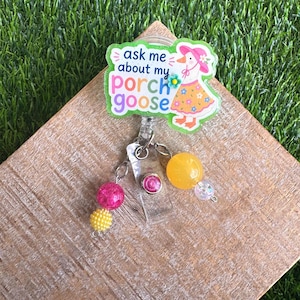 May include: A badge reel with a cartoon goose wearing a hat and dress. The badge reel has the text "ask me about my porch goose". The reel has pink and yellow beads and a yellow bead.