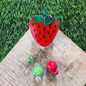 May include: A red glittery strawberry-shaped badge reel with black seed details and a green glitter top. The badge reel has a silver clip and a charm with green and red beads.