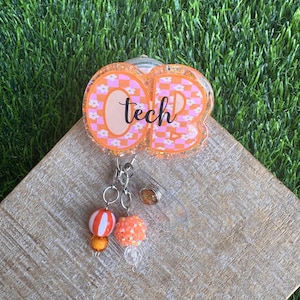 May include: An orange and pink badge reel with the letters "OB" and the word "tech" in a script font. The letters have a checkered pattern with small white flowers. The reel has a clear plastic clip and decorative charms.
