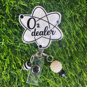 A white and black badge reel with the text "O2 dealer" in a science-themed design. The badge reel has a clear plastic clip and is adorned with black and white striped and pearl-like beads.