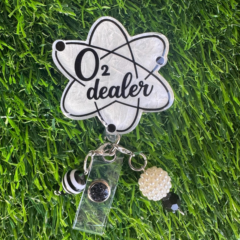 A white and black badge reel with the text "O2 dealer" in a science-themed design. The badge reel has a clear plastic clip and is adorned with black and white striped and pearl-like beads.