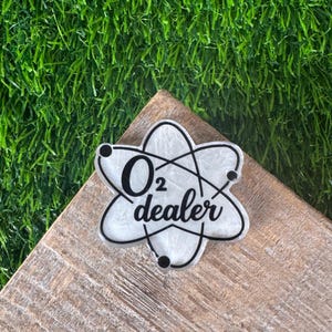 A white and black O2 dealer badge in the shape of an atom. The badge is on a wooden surface with a green, artificial grass background.