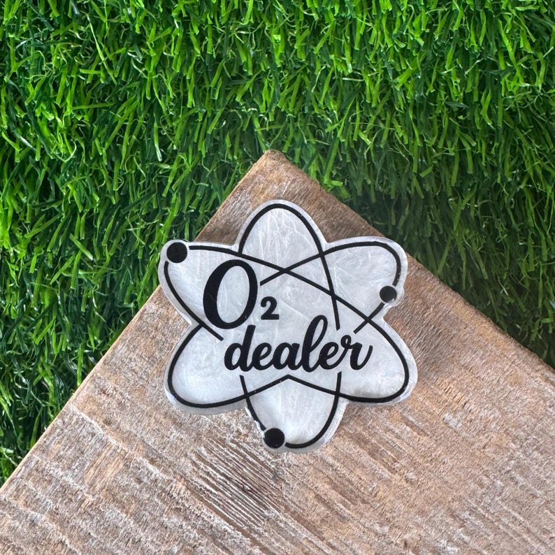 A white and black O2 dealer badge in the shape of an atom. The badge is on a wooden surface with a green, artificial grass background.