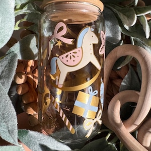 May include: A glass jar with a wooden lid decorated with a rocking horse, a candy cane, a gift box, and a bow. The jar is filled with a white substance.